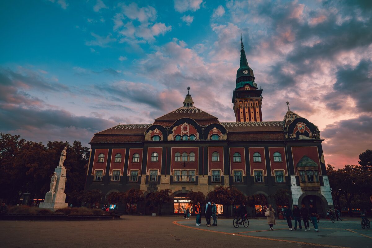 Subotica City Hall, red with ornate gables and a green-topped tower, seen across the square at dusk under pink clouds