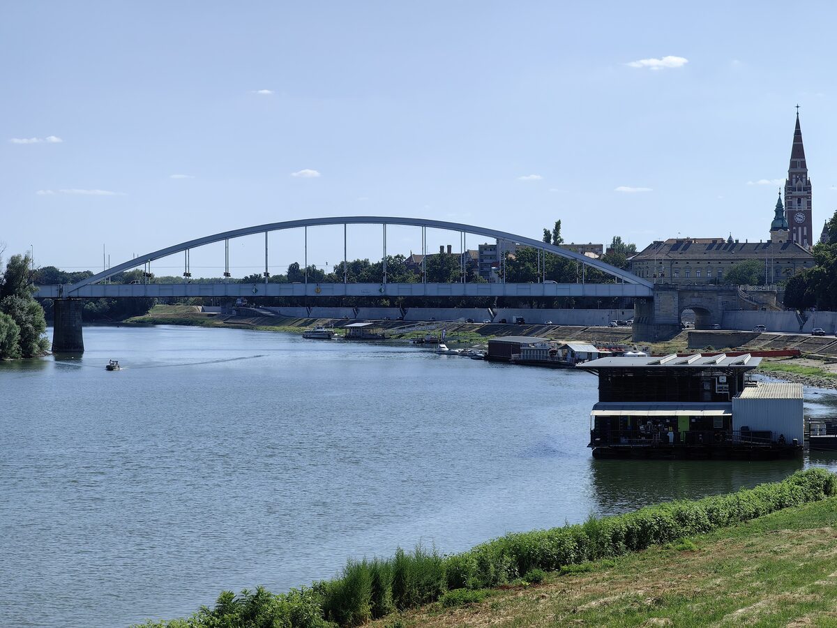 The Tisza flowing through Szeged under the arched steel Belvárosi Bridge, with a floating pontoon and a church spire on the right bank