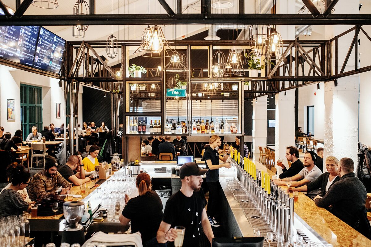 The long taps and bar counter of the Põhjala Tap Room, with guests at the bar under industrial lamps