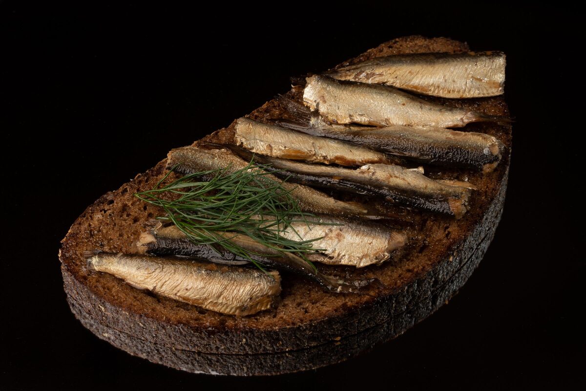 A slice of dark rye bread topped with sprats and a sprig of dill on a black background