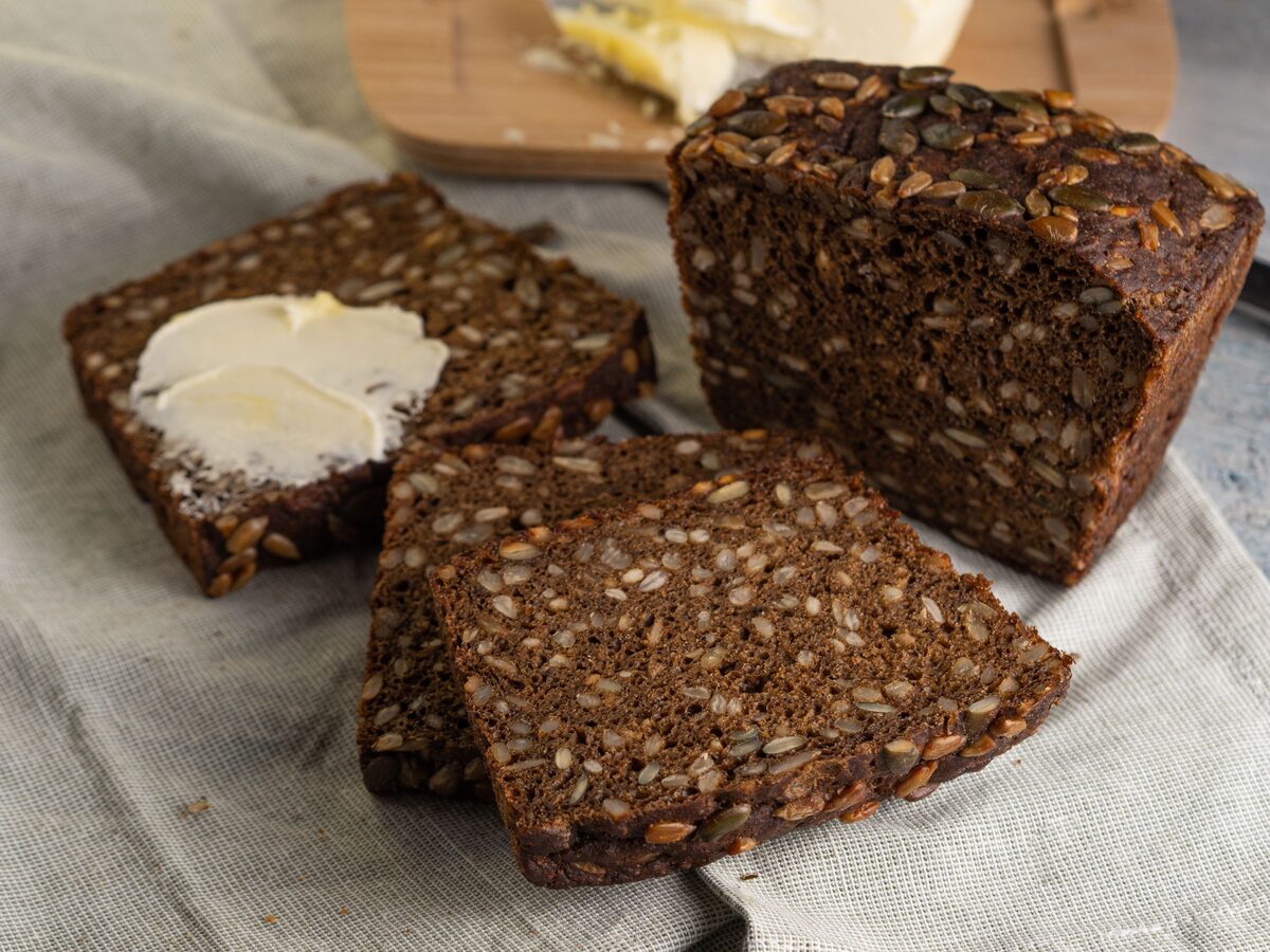 Slices and a loaf of dense dark rye bread with seeds, one slice spread with butter
