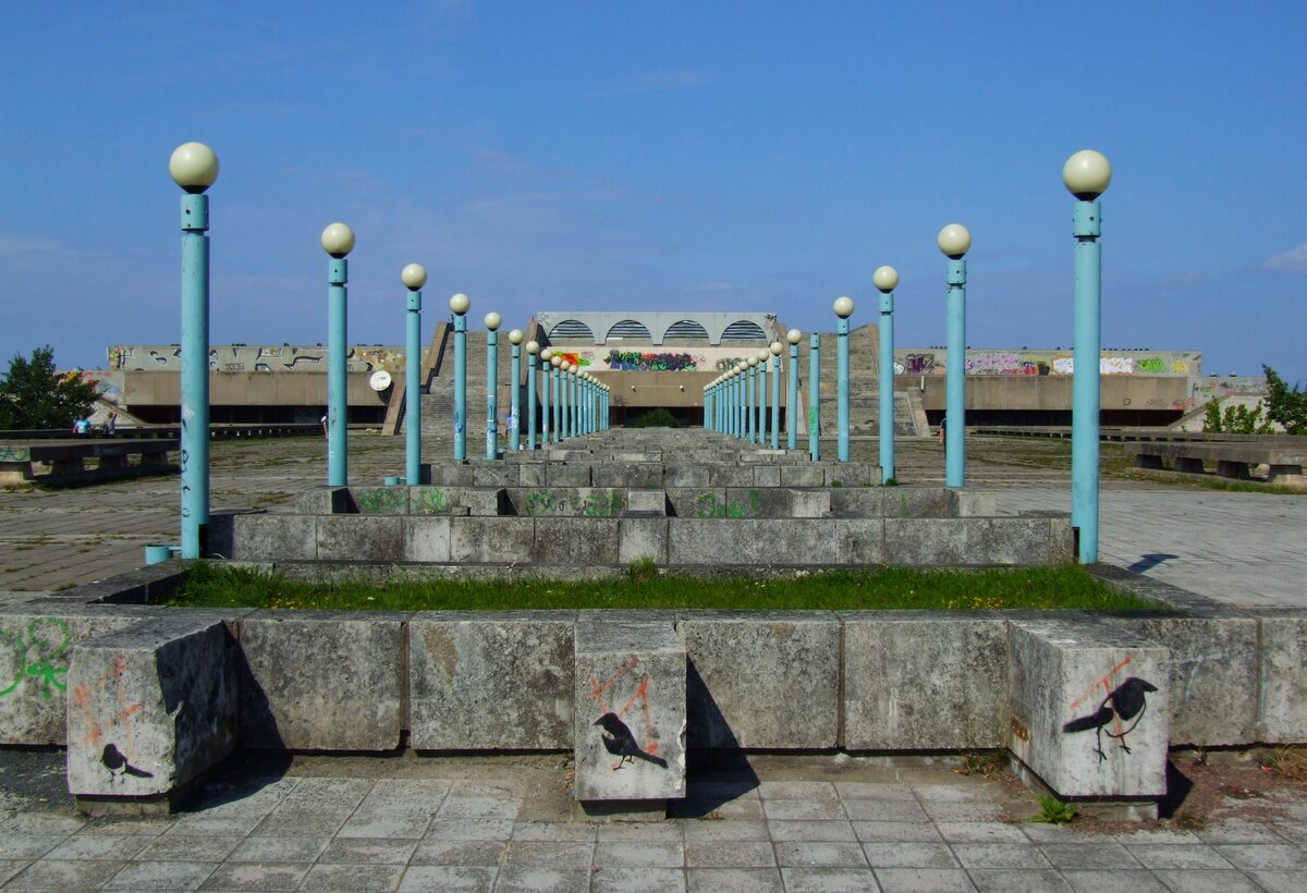 A row of tall blue-green lamp posts and low concrete steps on the Linnahall terrace, with concrete blocks painted with birds in front