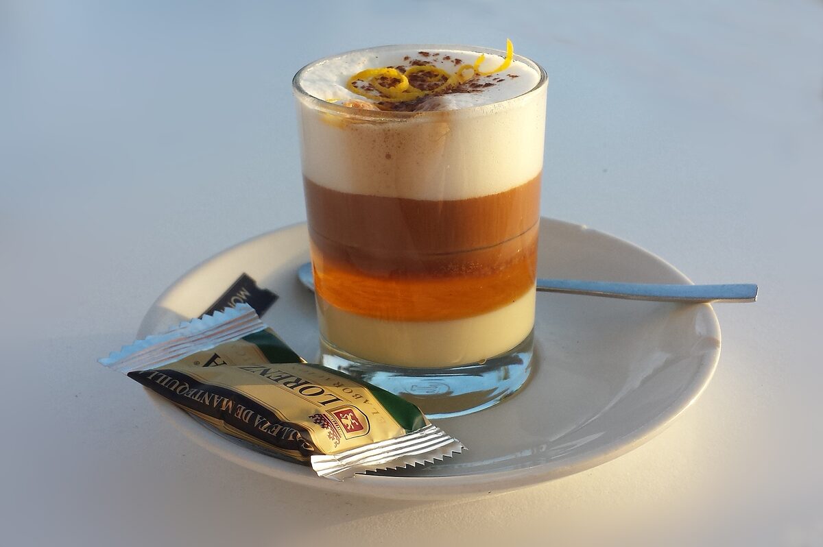 A layered barraquito in a glass, with a lemon peel and a sugar sachet beside it