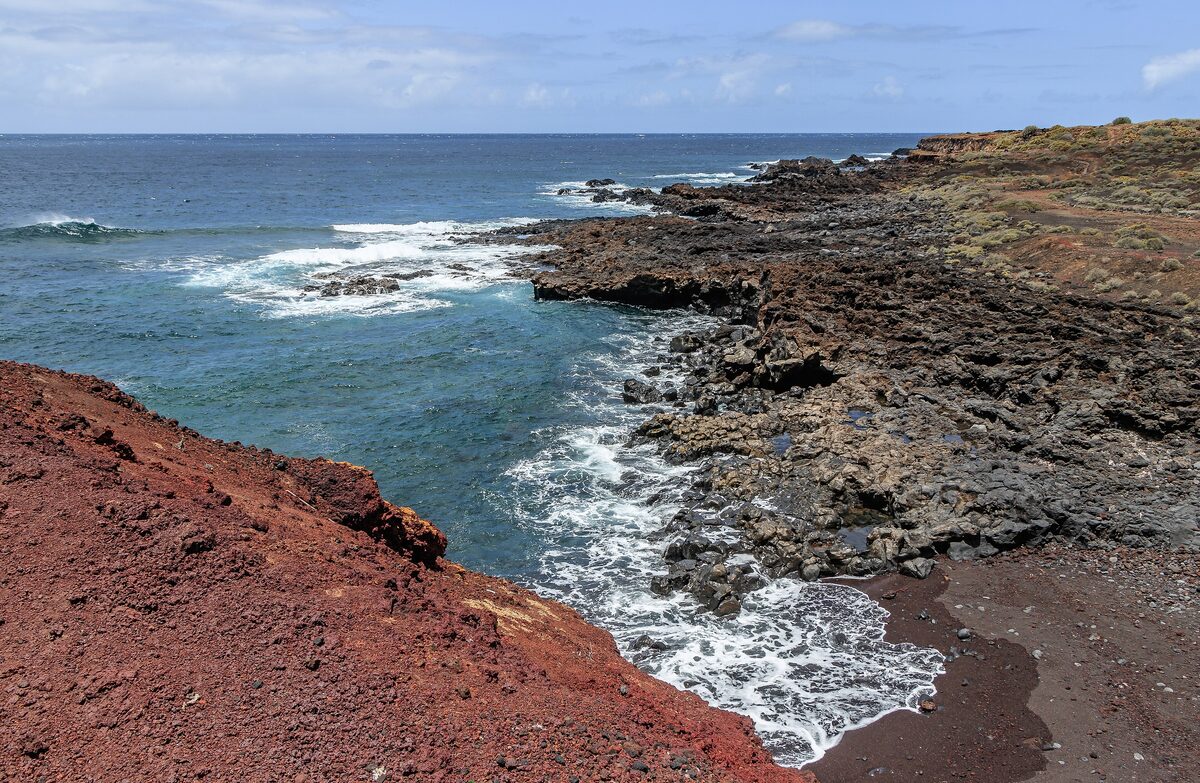 A red volcanic hillside and black lava shore with surf at Punta de Teno