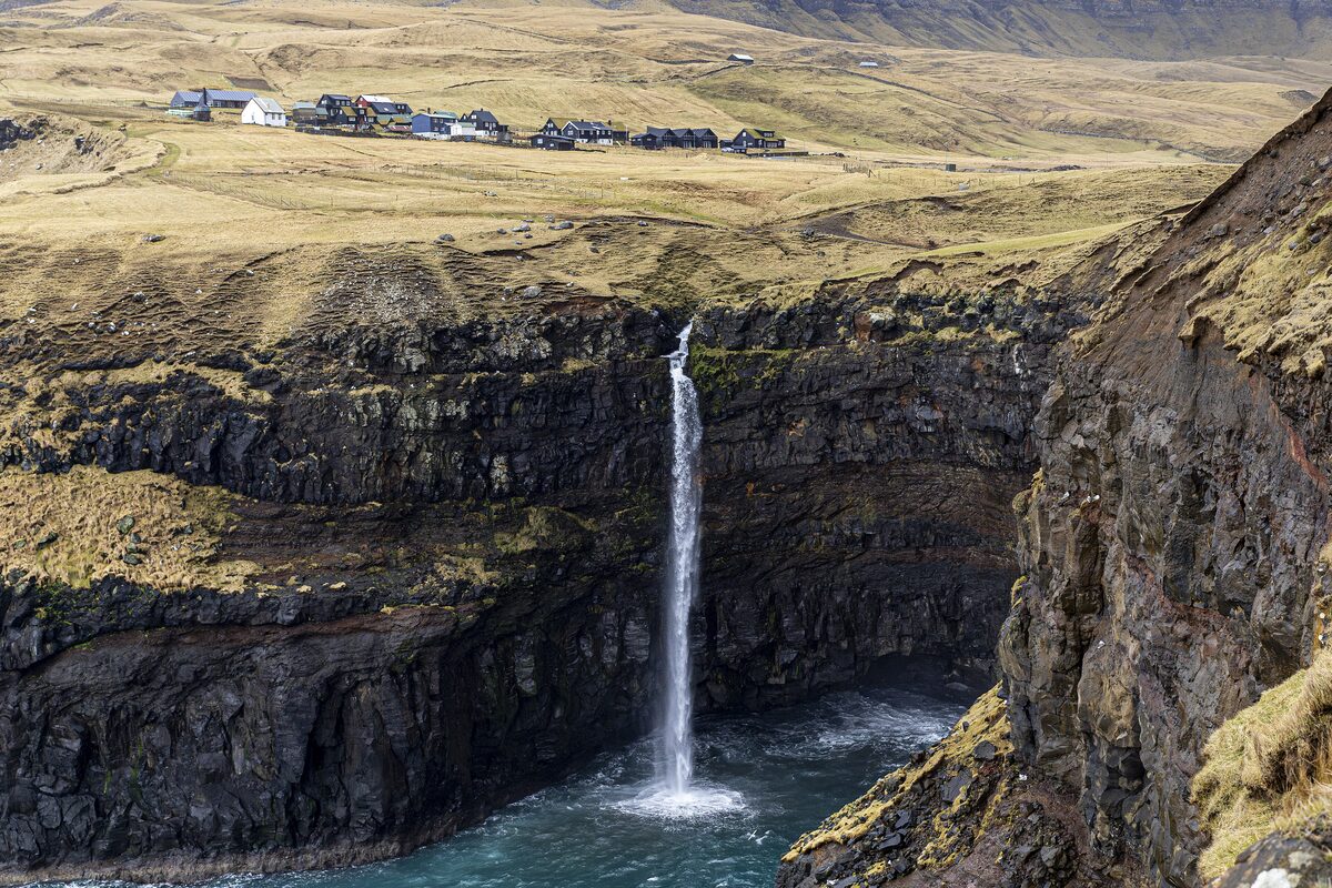 The houses of Gásadalur on the plateau above the cliffs, with the Múlafossur waterfall dropping into the sea below