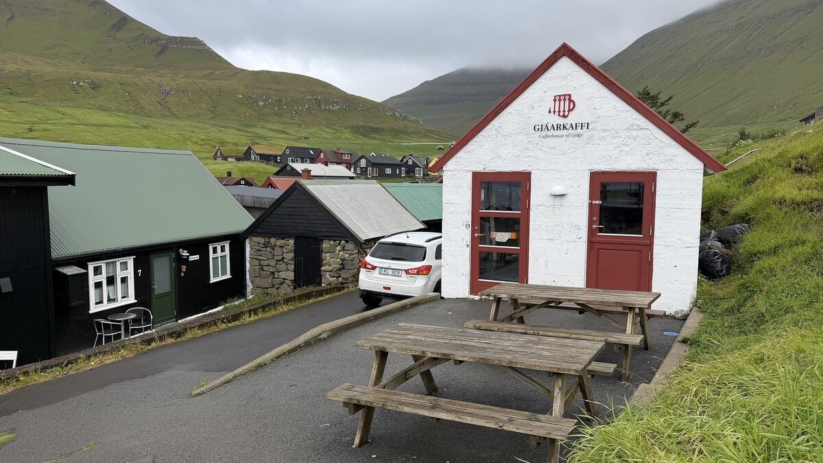 The small white Gjáarkaffi coffee house with red doors and picnic tables, with green mountains behind Gjógv