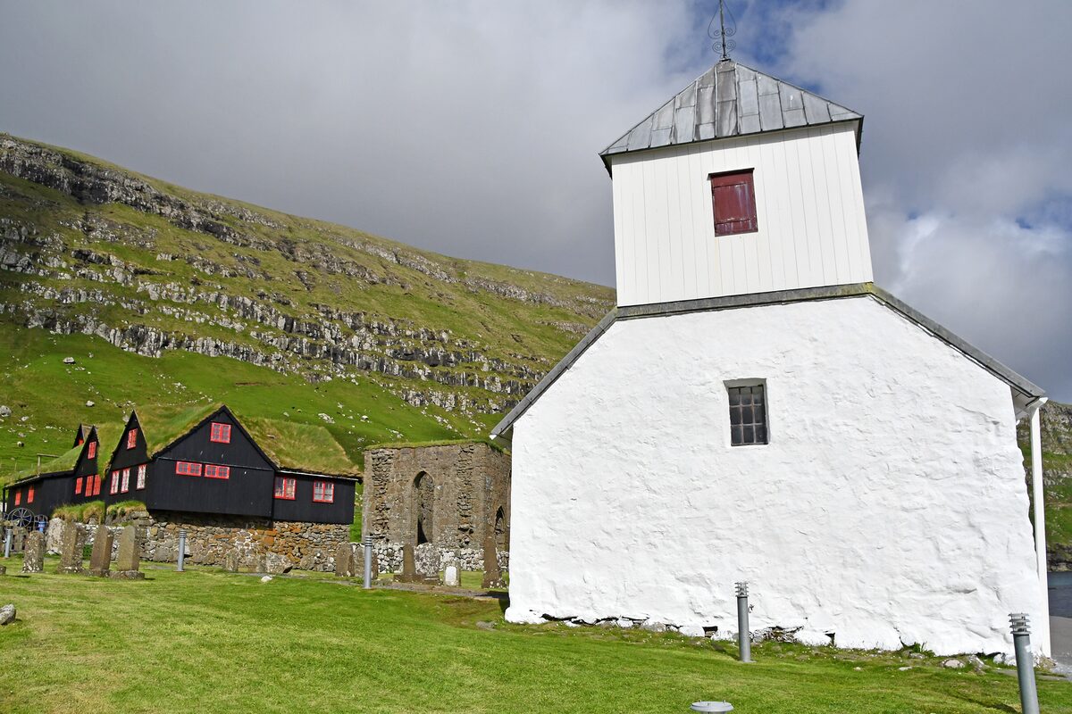 The whitewashed St. Olav's Church in Kirkjubøur with the roofless Magnus Cathedral walls and black farm buildings behind it