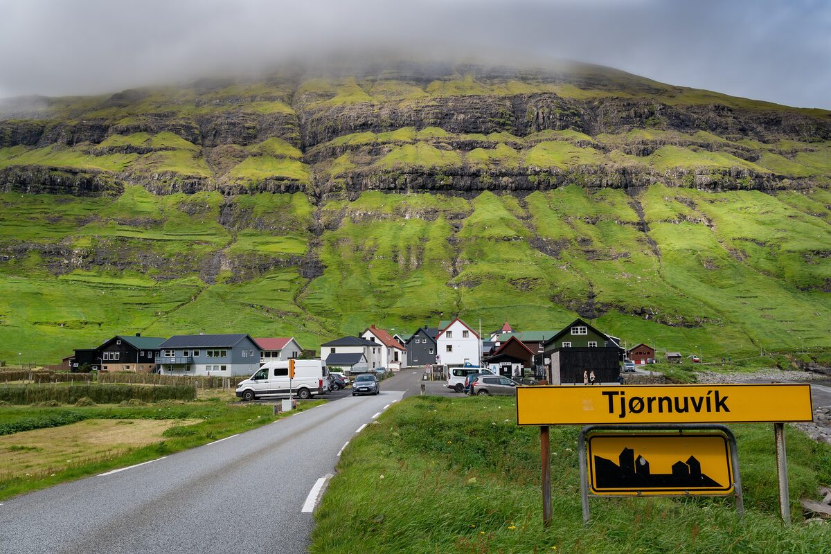 The village of Tjørnuvík under a cloud-capped green cliff, with a village sign in the foreground