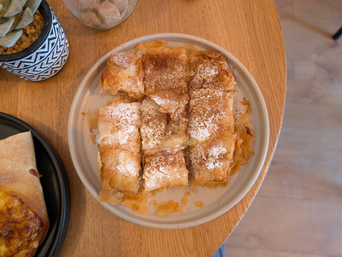 A plate of bougatsa cut into squares and dusted with powdered sugar and cinnamon on a wooden café table