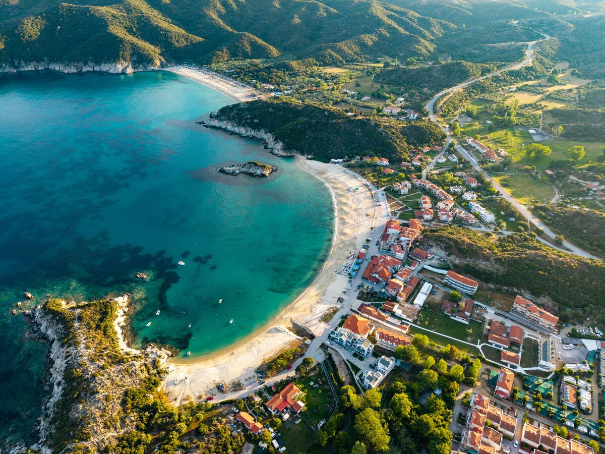 Aerial view of a curved beach in Sithonia with turquoise water, a small island in the bay and a village along the shore