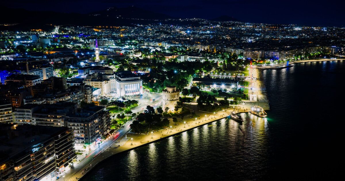 Thessaloniki at night from the air, with the lit White Tower on the waterfront promenade, the city spreading behind it and the dark sea on the right