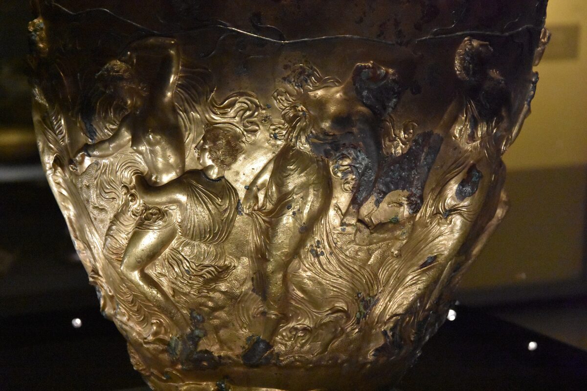 Close-up of the gilded bronze Derveni krater, a wine vessel covered in relief figures of Dionysus and his followers, glowing in a dark museum room