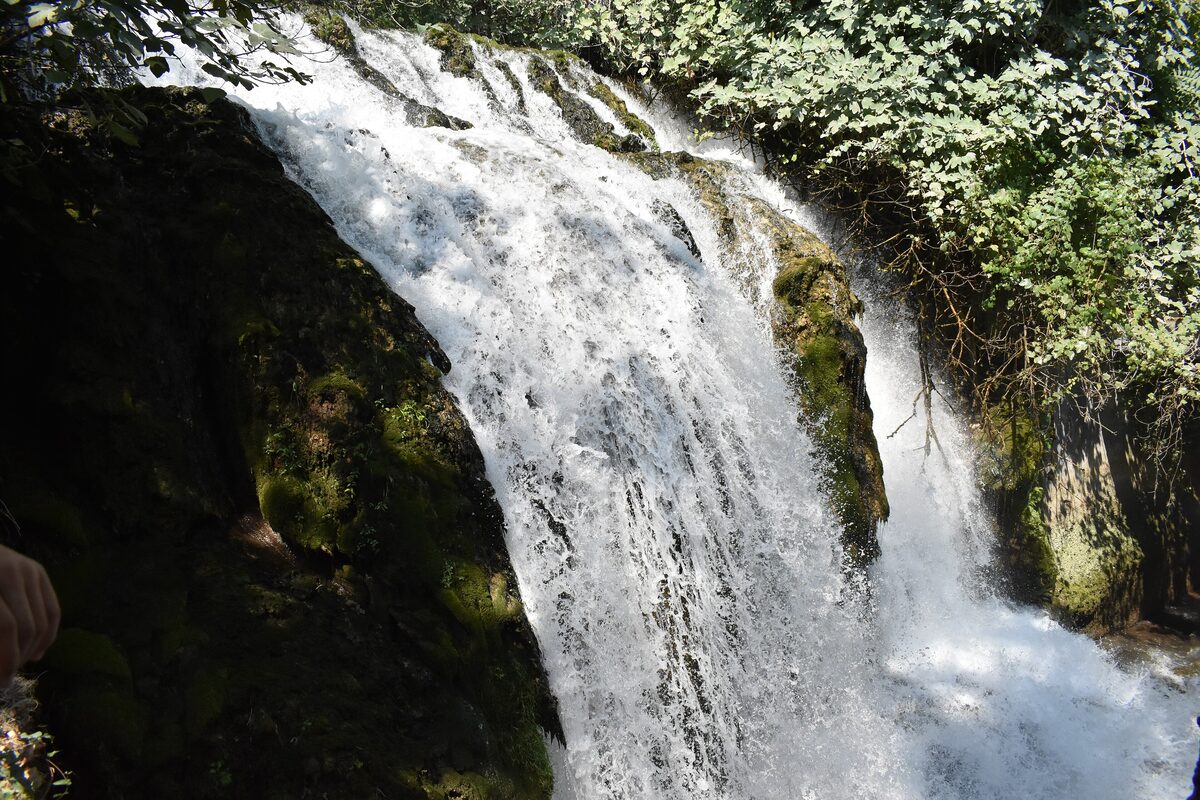 A waterfall at Edessa cascading down a dark, moss-covered cliff surrounded by green trees