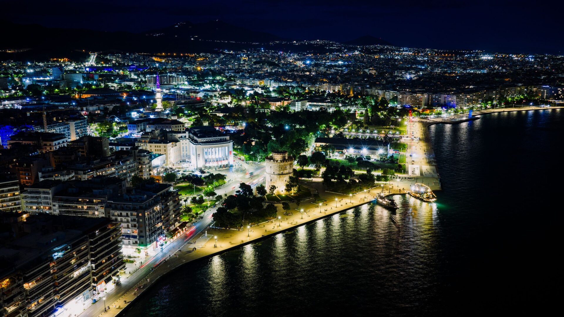 Thessaloniki at night from the air, with the lit White Tower on the waterfront promenade, the city spreading behind it and the dark sea on the right