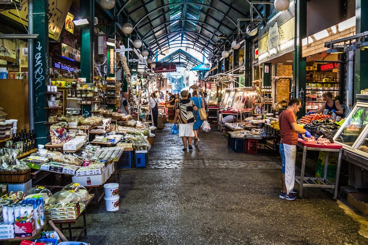 A covered aisle of Kapani market with stalls of vegetables, olives and fish on both sides under an arched iron roof