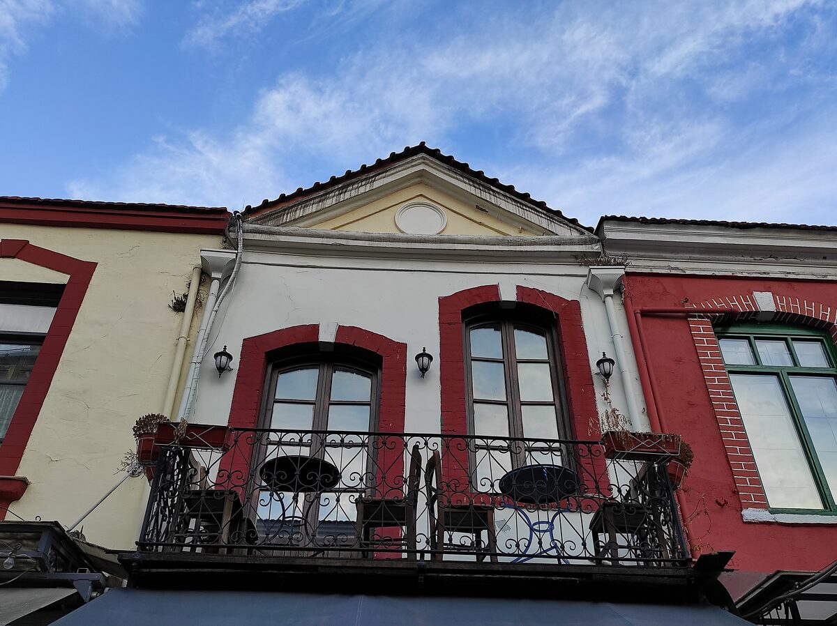 A pastel-painted old building in Ladadika with red window frames and a wrought-iron balcony set with café tables