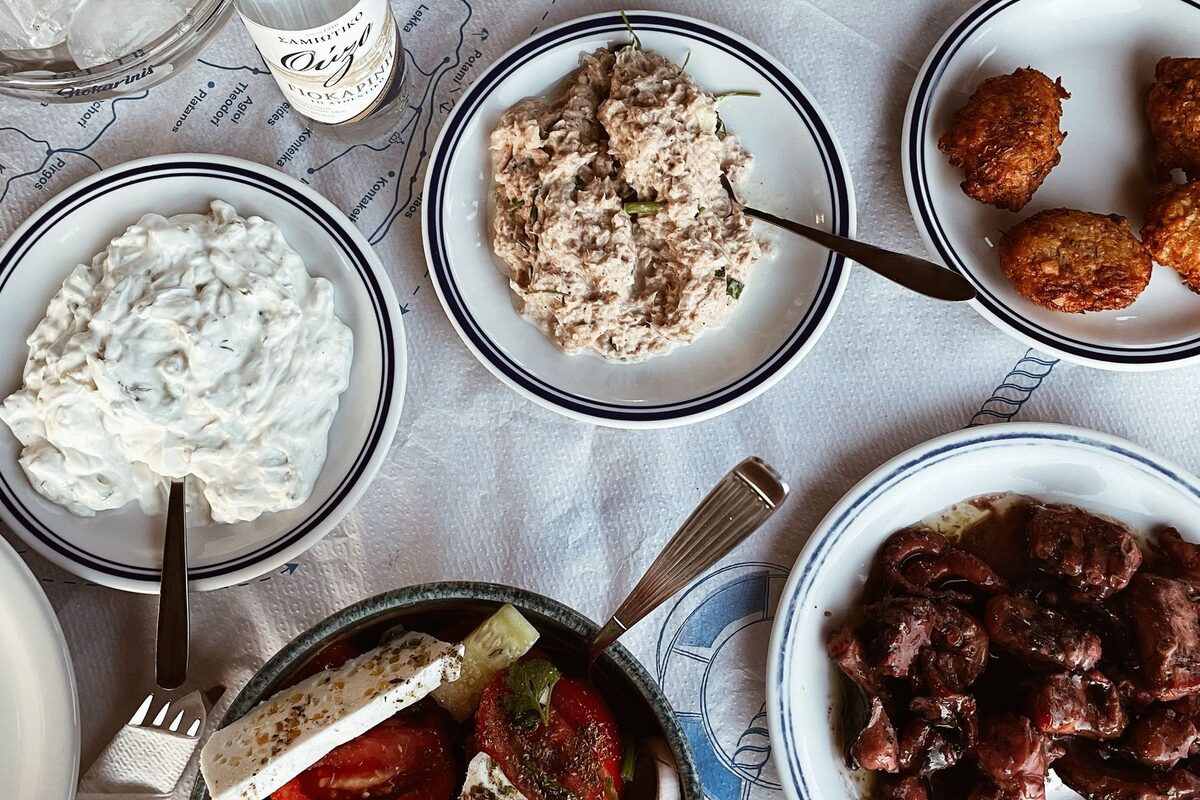 A table of Greek meze seen from above: tzatziki, fish roe dip, fritters, octopus in red wine and a tomato salad with a slab of feta, with a bottle of ouzo