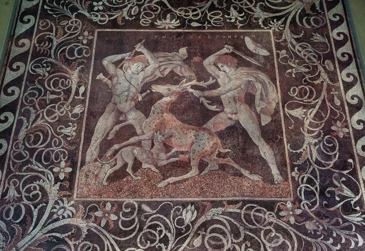 An ancient pebble mosaic from Pella showing two hunters with a sword and an axe attacking a stag, inside a floral border