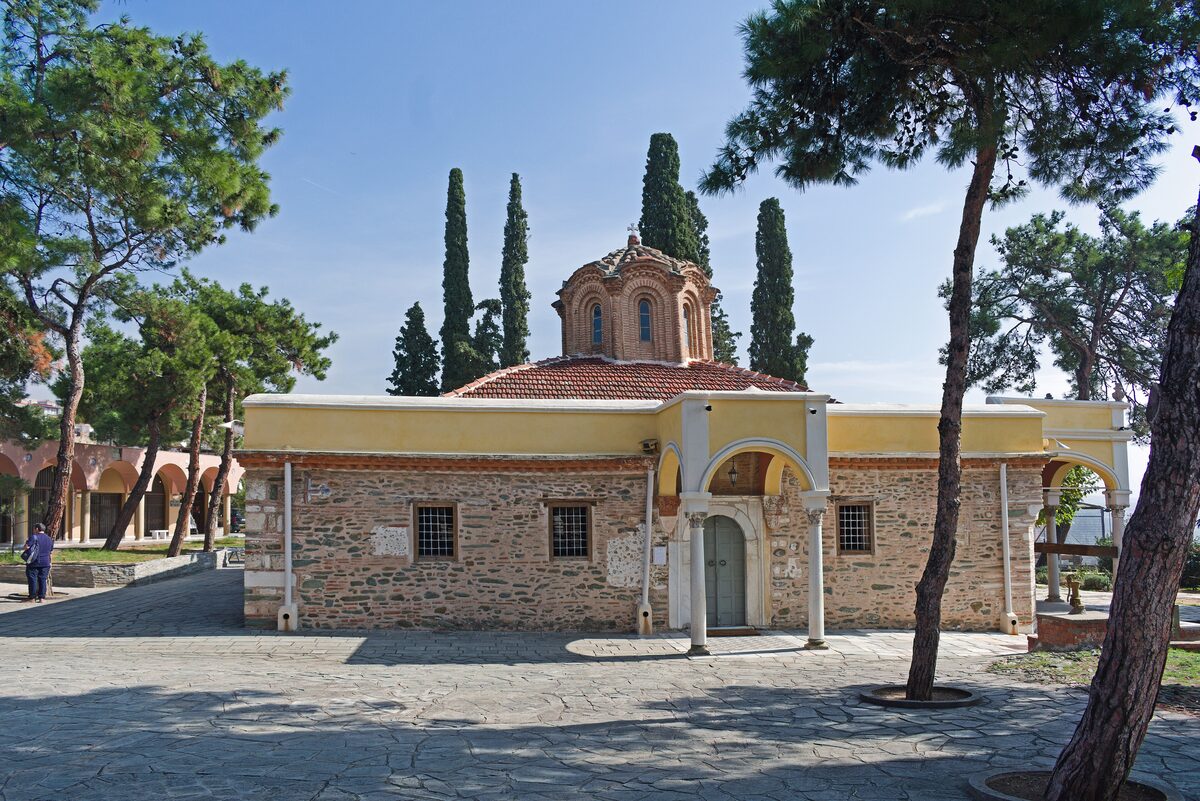 The small Byzantine main church of the Vlatadon Monastery with its brick dome, framed by pine and cypress trees in a paved courtyard