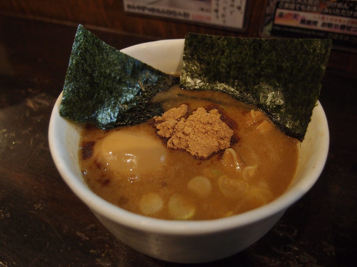 A bowl of thick dipping broth with a half egg, nori and fish powder, the cold noodles on a plate behind it, at the Fuunji counter in Shinjuku