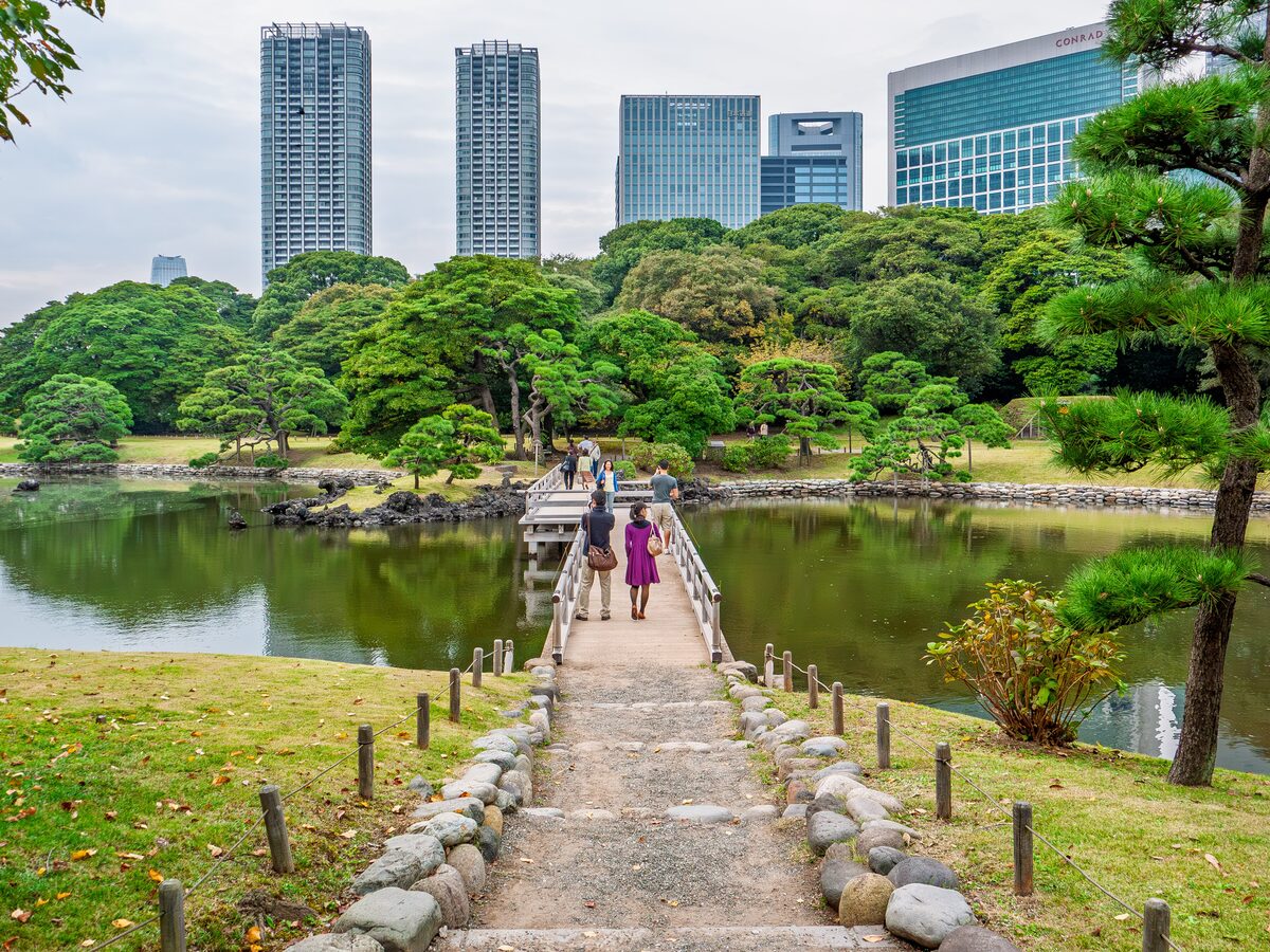 A stone-edged path and wooden bridge crossing a pond in Hamarikyu Gardens, with office towers behind the trees