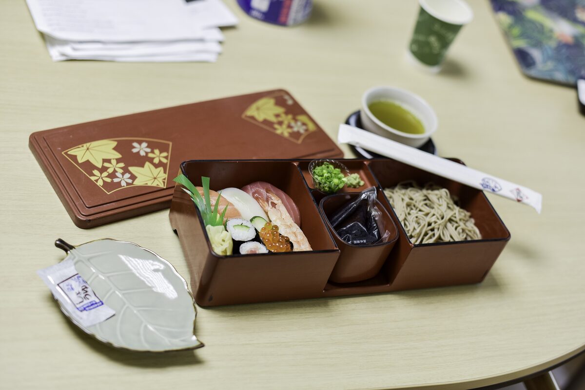 A bento box with nigiri sushi, soba noodles and pickles beside a cup of green tea on a table