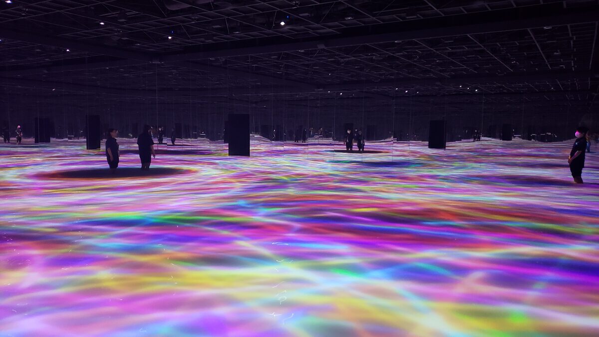 A dark teamLab Planets hall with a floor of flowing, rainbow-coloured light and a few visitors standing in it