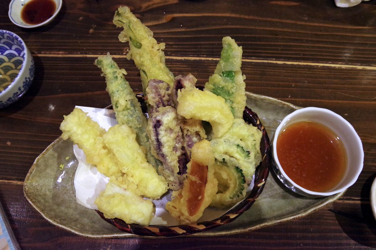 Vegetable tempura with a crisp golden batter on a plate, with a bowl of dipping sauce