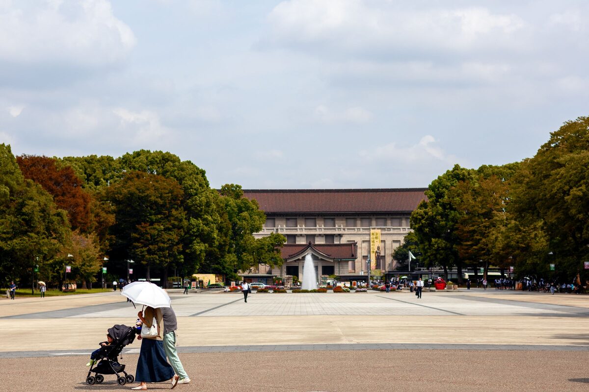 The long front of the Tokyo National Museum seen across the open plaza in Ueno Park, with trees on both sides and a pair of walkers in the foreground