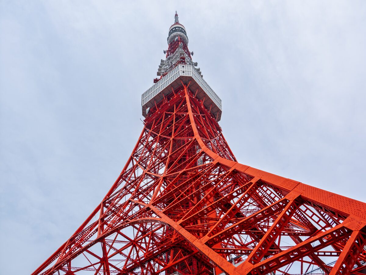 Looking up at Tokyo Tower's red-and-white steel lattice rising to the main deck and antenna under a pale sky