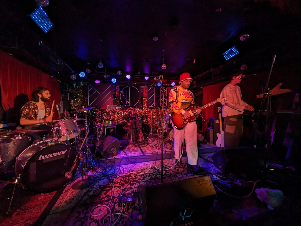 A band playing on the small stage of the Horseshoe Tavern under colored lights, with a red curtain and a drum kit