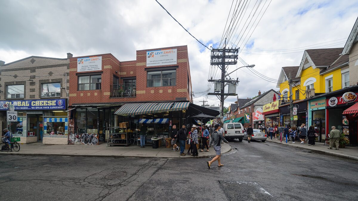 A Kensington Market street corner with cheese and produce shops, sidewalk stalls and yellow-painted houses