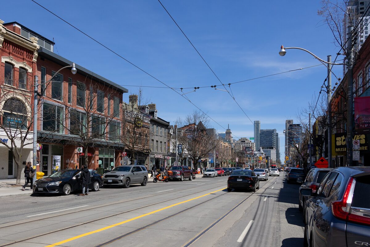 Queen Street West looking east along the streetcar tracks, with red-brick shopfronts on the left and the downtown towers ahead