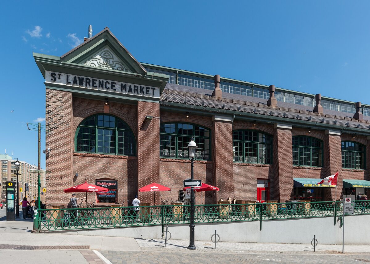 The brick South Market building of St. Lawrence Market with its gabled entrance and red patio umbrellas under a blue sky
