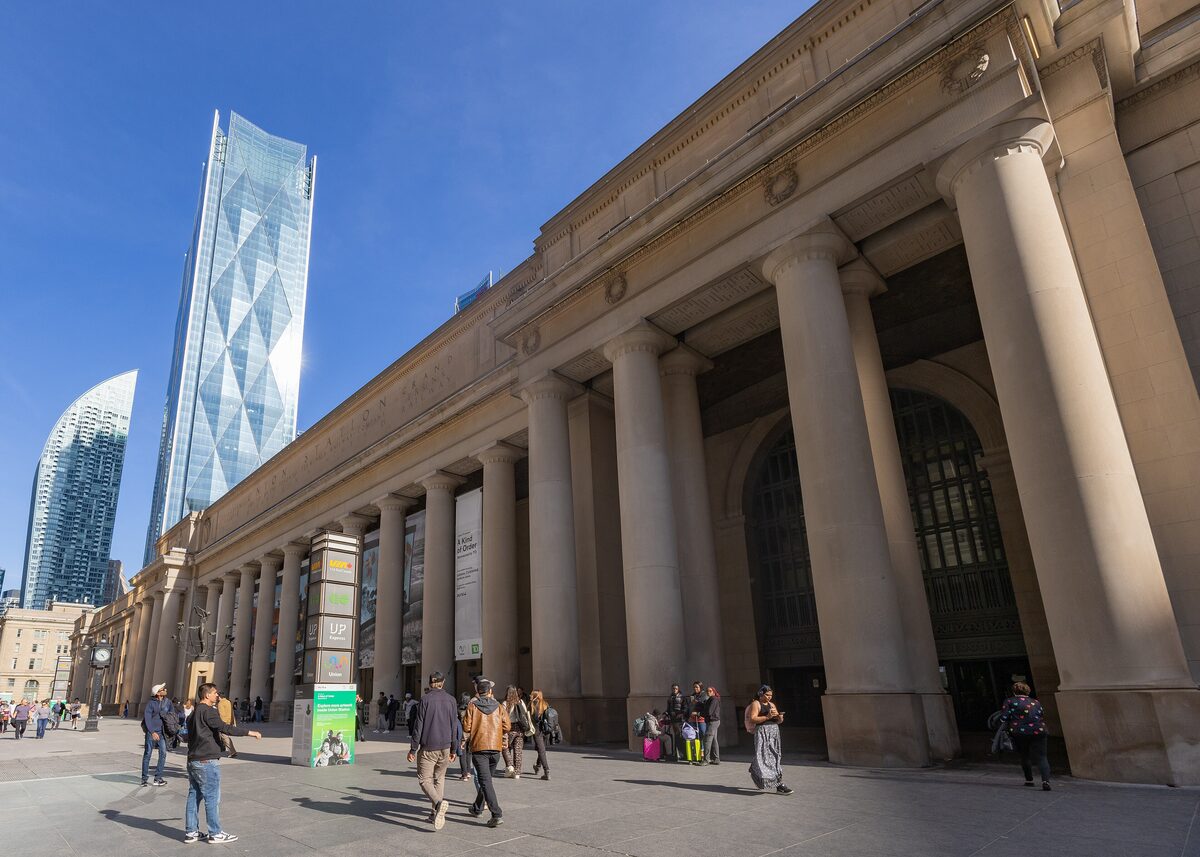 The stone colonnade of Union Station's Front Street facade in afternoon sun, with glass towers behind and people walking past