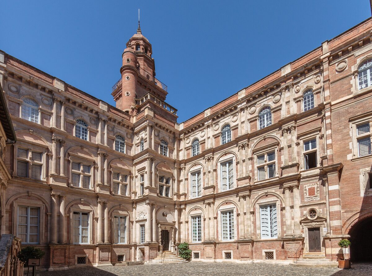 The courtyard of the Hôtel d'Assézat in Toulouse, with brick and stone walls, tiered window frames and a tall tower under a blue sky