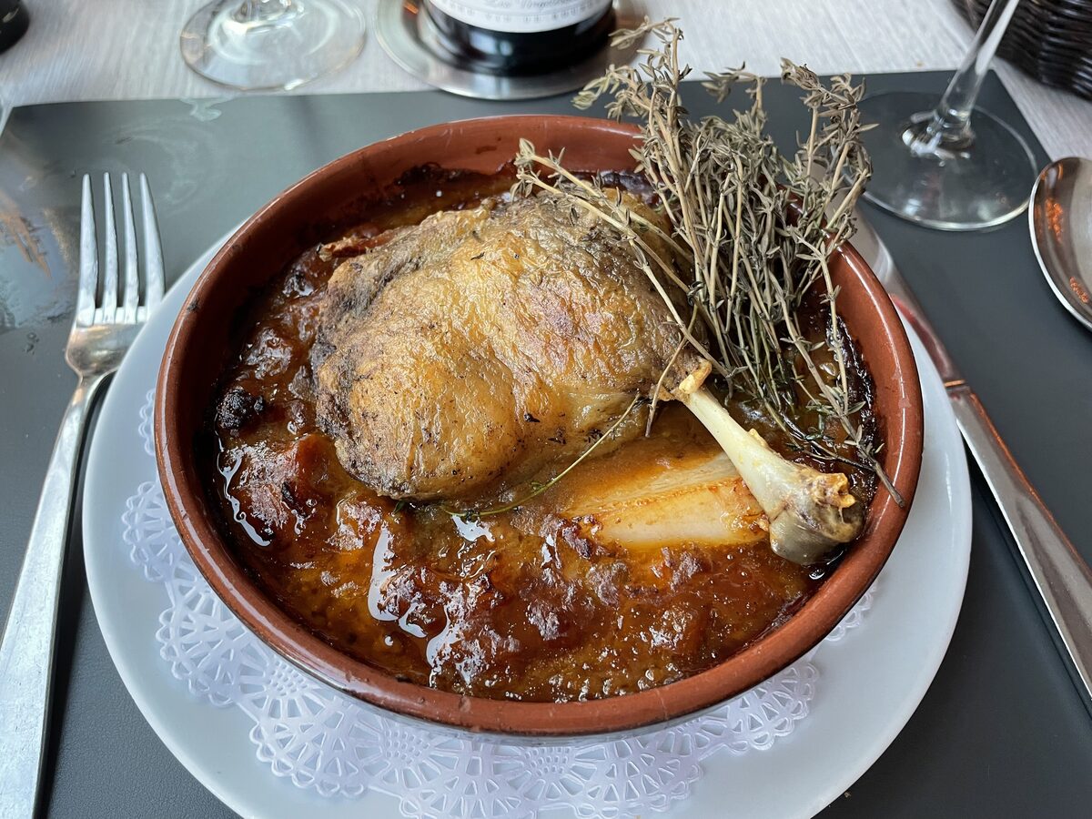 A clay dish of cassoulet topped with a duck leg, set on a table with cutlery