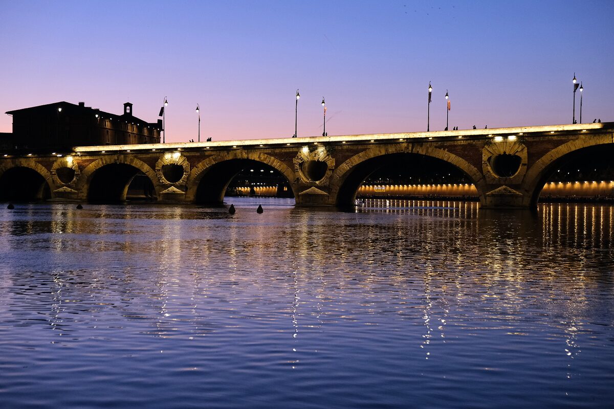 The Pont-Neuf over the Garonne at dusk, its arches lit from below and reflected in the water under a purple-pink sky
