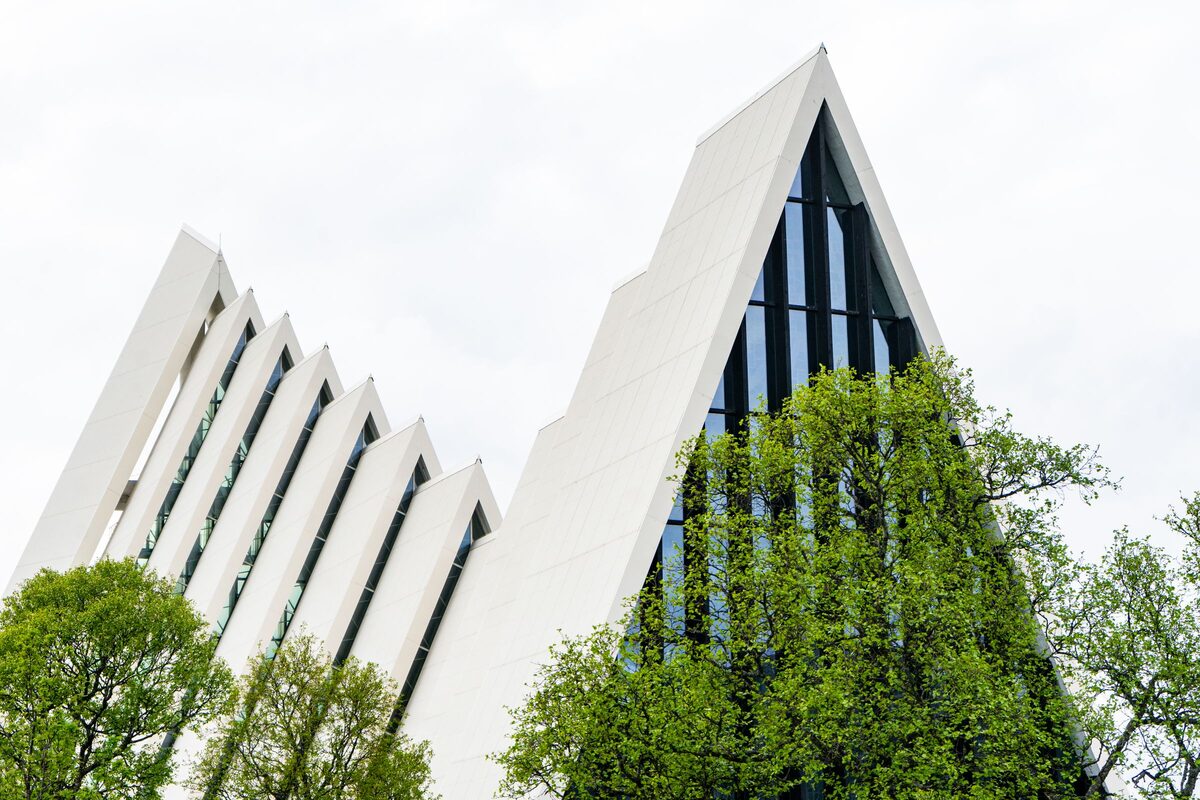 The white, stepped triangular roofs of the Arctic Cathedral in Tromsø with green trees in front