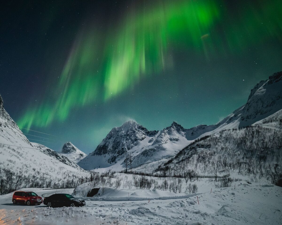 Green northern lights rippling across a starry sky above snowy mountains and two parked cars