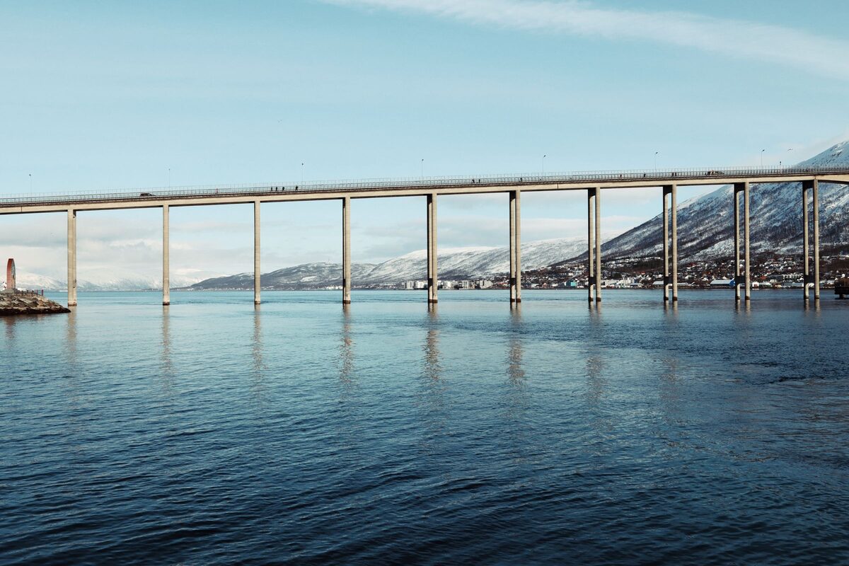 The long, high Tromsø Bridge on slender pillars over calm water, with snowy mountains behind
