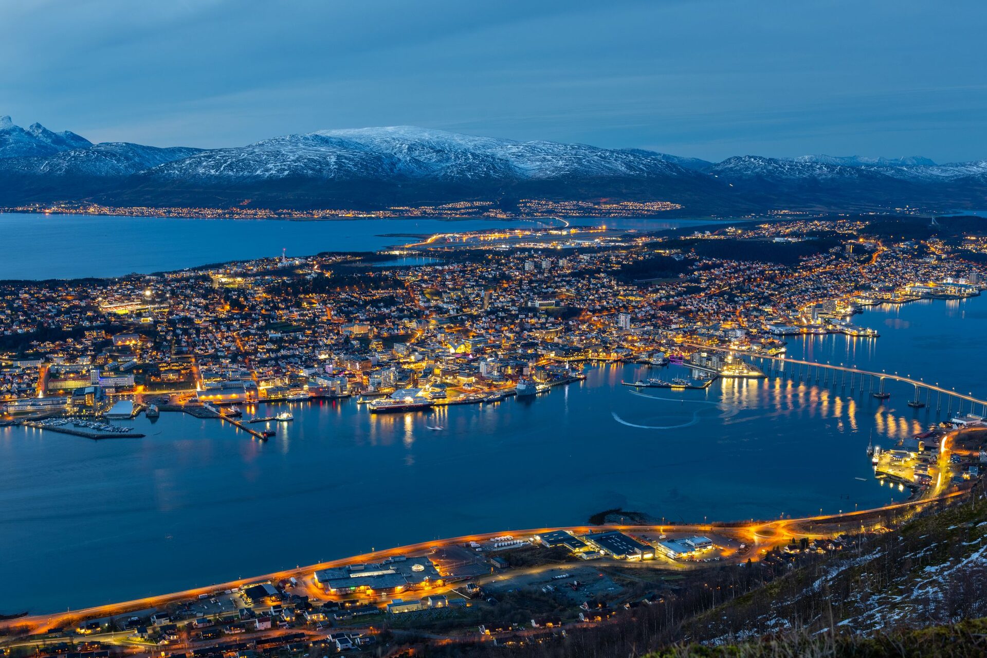 Tromsø at blue hour seen from above, with the lights of the island city, the long bridge to the mainland and snowy mountains across the fjord