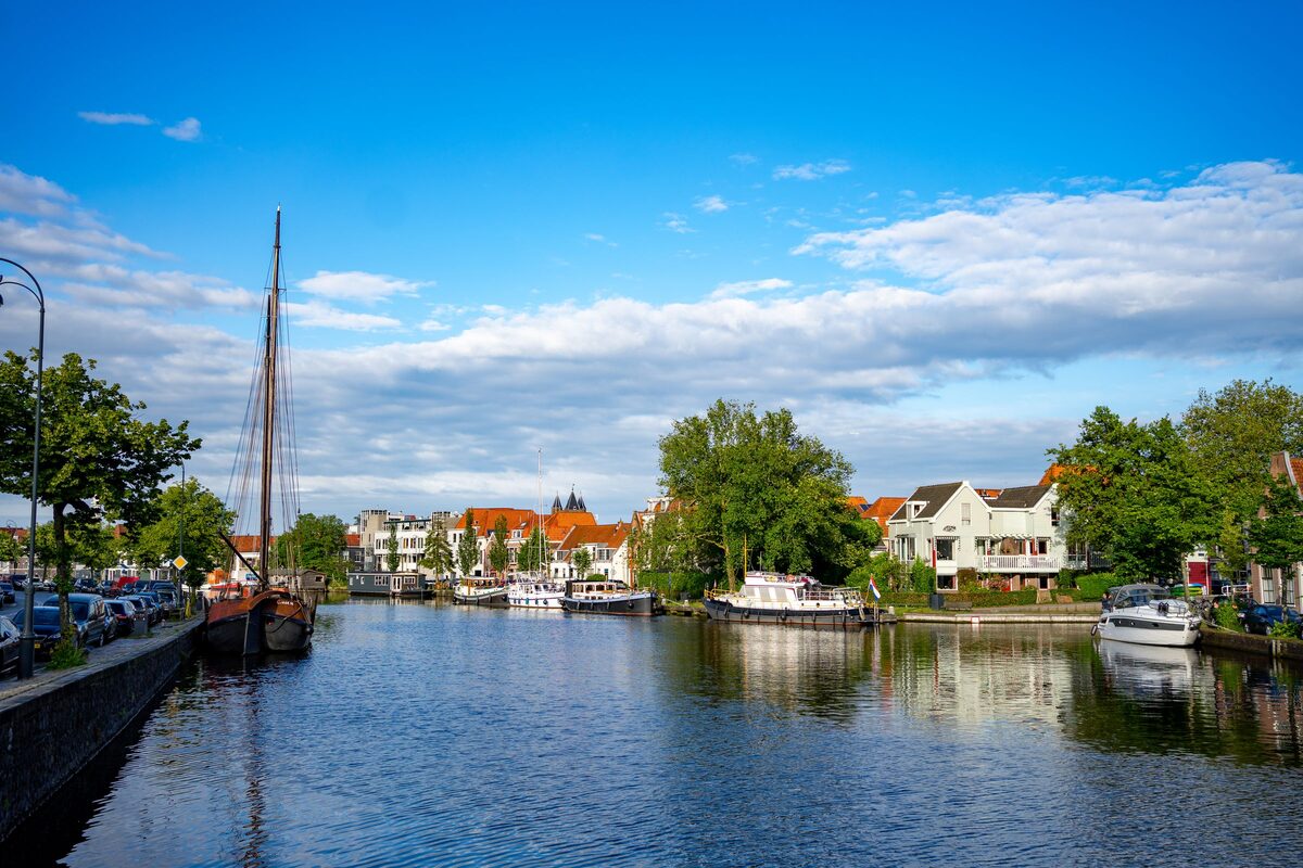 A wide canal in Haarlem with an old sailing ship, moored boats and houses among trees under a blue sky