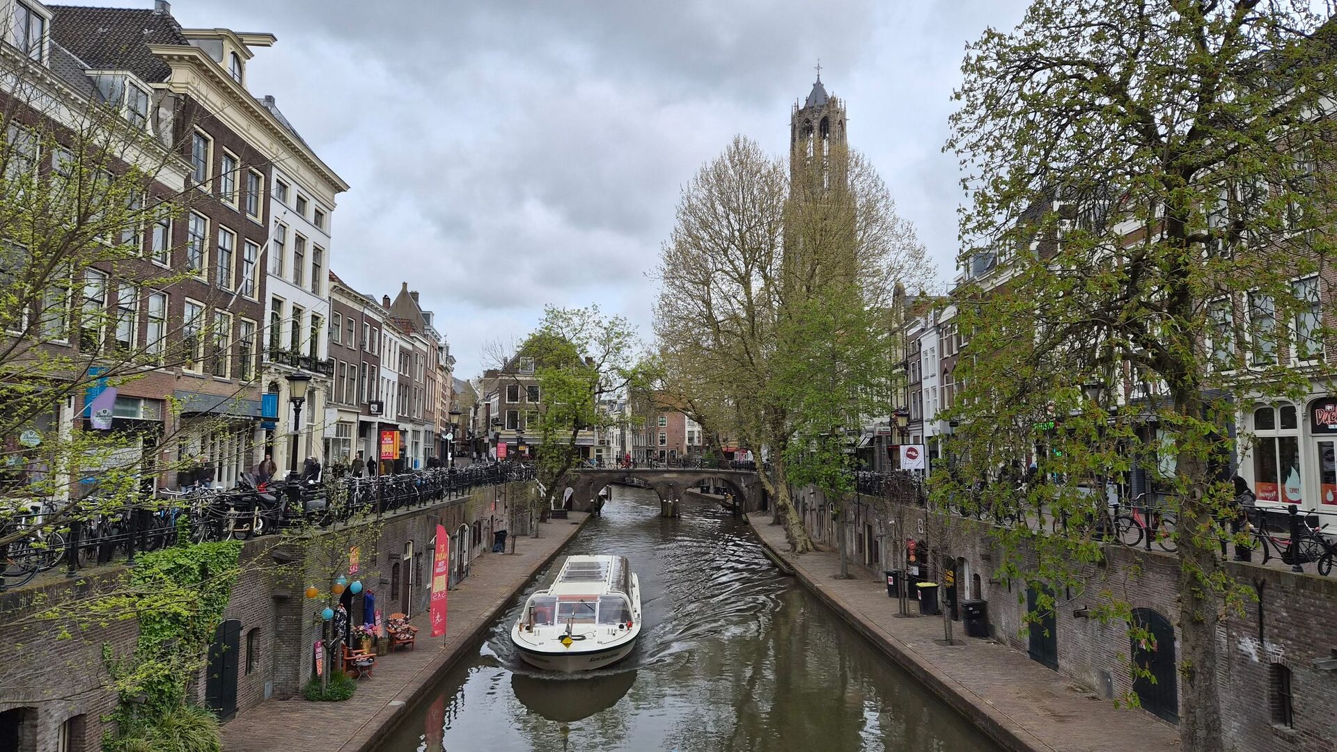 A tour boat on the Oudegracht in Utrecht, with the wharves and cellars at water level, old houses, spring trees and the Dom Tower at the end of the canal