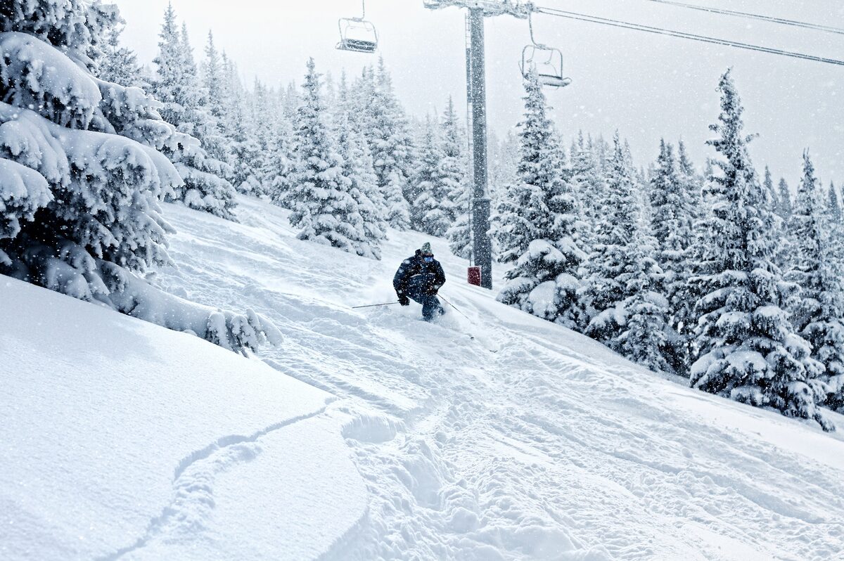 A skier in deep powder below a chairlift, with snow-covered spruce trees lining the run