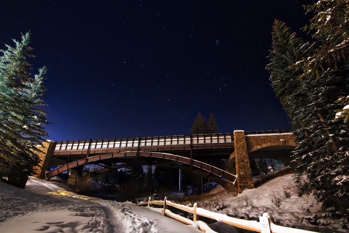 The covered bridge in Vail Village at night, with snow on the banks and stars over the trees