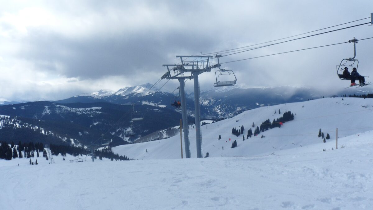 A chairlift carrying skiers over a wide snowy slope on Vail Mountain, with distant ridges under a cloudy sky