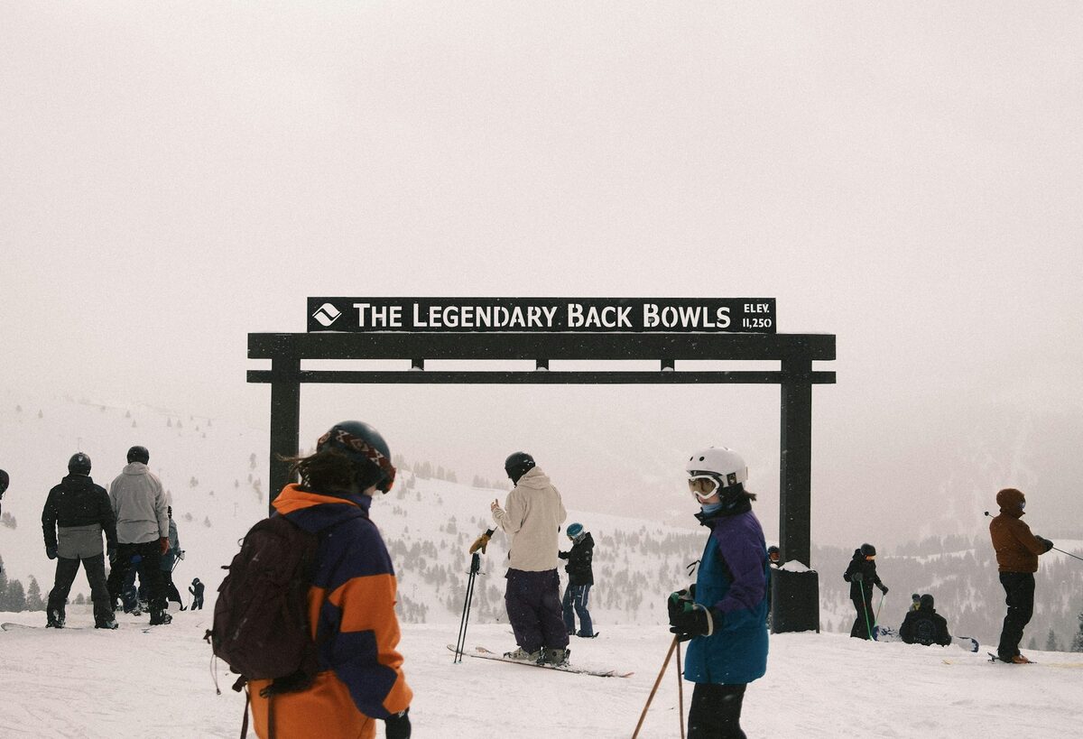 Skiers gathered under the wooden Legendary Back Bowls entrance sign on a snowy, hazy ridge