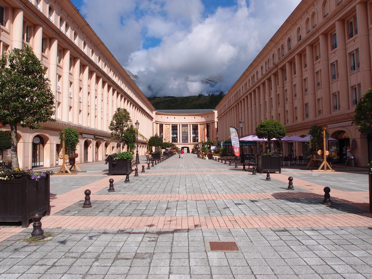 The arcaded square of Albertville, with pale stone buildings, bollards and café terraces under a cloudy sky