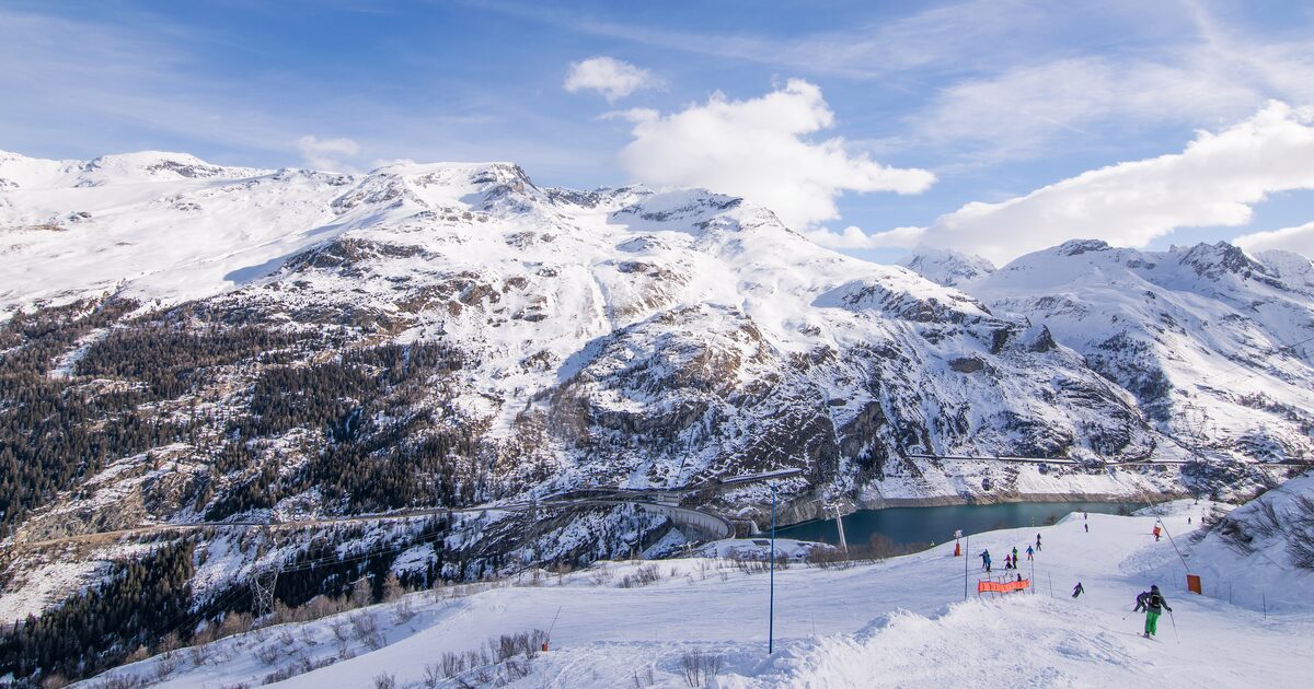 Skiers on a snowy piste above the Chevril lake and the Tignes dam, with snow-covered mountains behind
